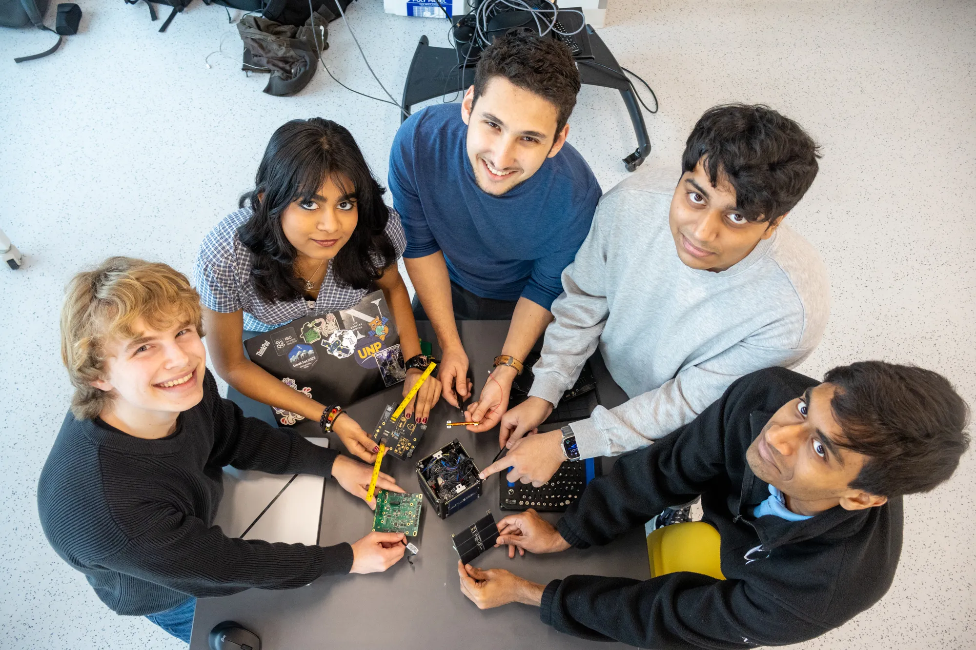 Five club members look up at the camera from around a table covered in CubeSat boards and tools
