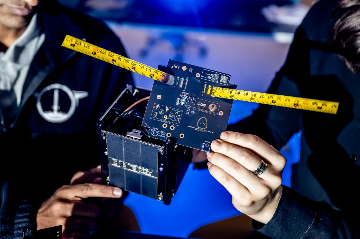 Two students hold a CubeSat and a black circuit board between them, with yellow tape measures extended